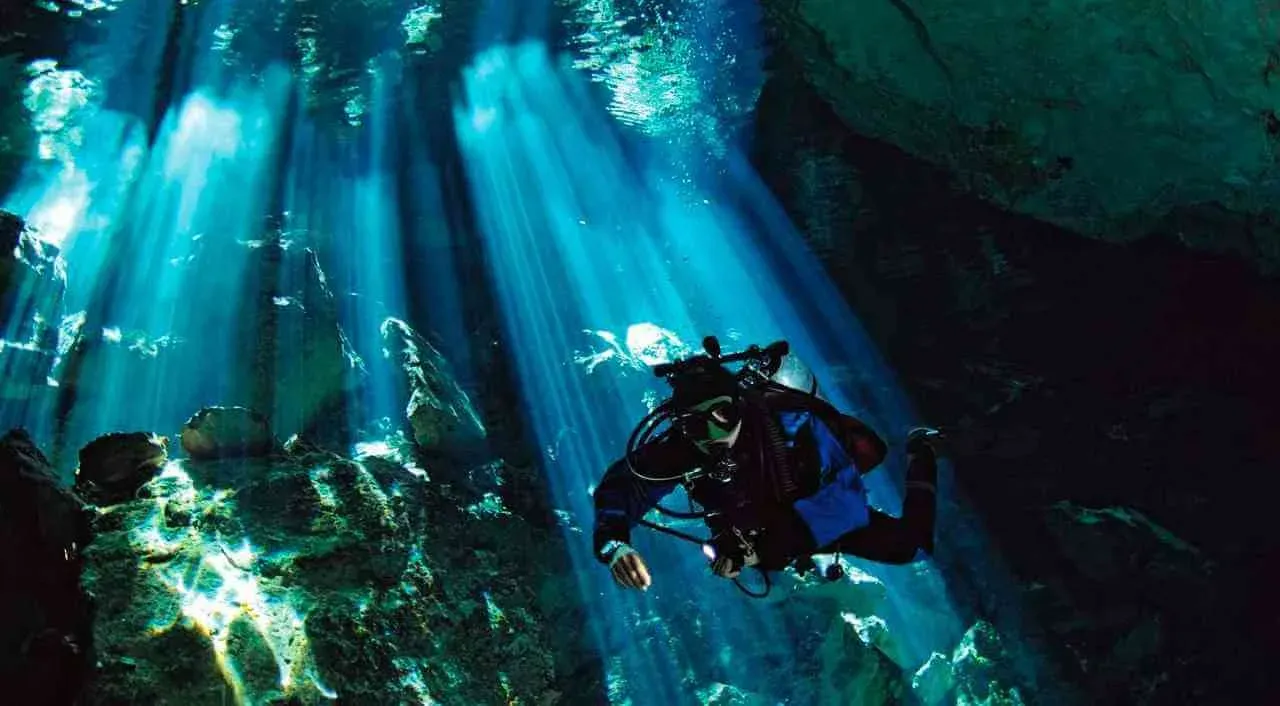 Mergulhador em caverna submersa atravessada por feixes de luz