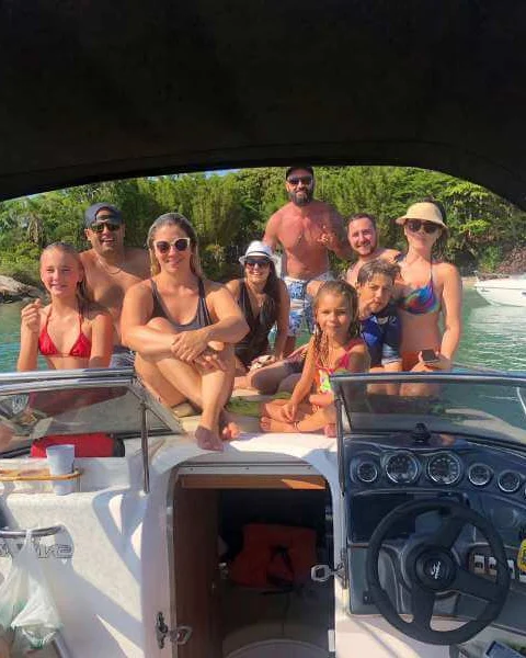 Família com crianças reunida na lancha, sorrindo para a foto diante de uma praia com mata