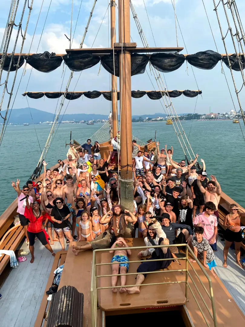 Convés do barco cheio de passageiros acenando, com os mastros e o mar ao fundo
