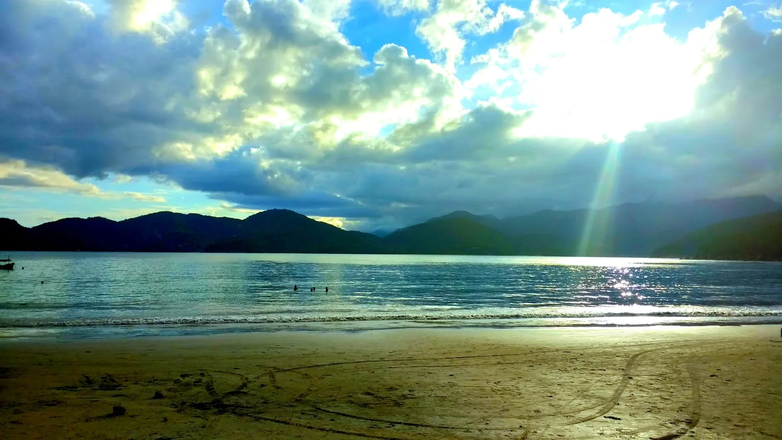 Banhistas na água rasa da Praia do Lázaro em Ubatuba, com a serra ao fundo e raios de sol atravessando as nuvens